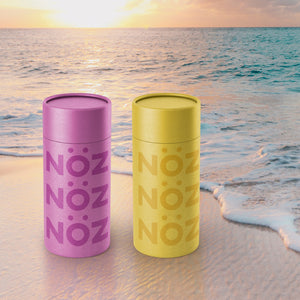 Two NOZ yellow and purple nose stick sunscreen in front to the beach.